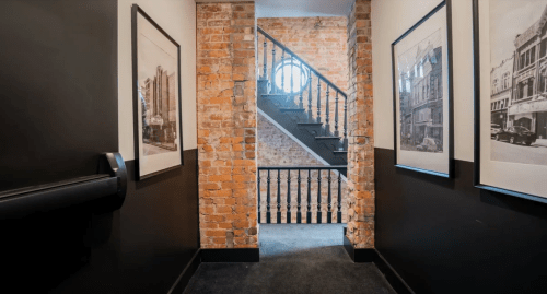 A hallway with exposed brick walls, framed black-and-white photos, and a staircase leading to the upper floor.