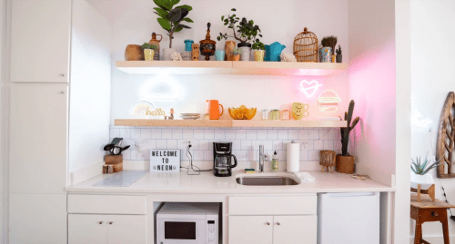 A bright kitchen with open shelves displaying plants, colorful dishes, and neon signs, featuring a coffee maker and microwave.