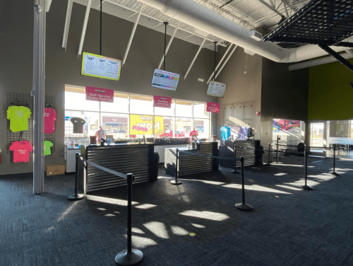 Brightly lit reception area with two counters, digital screens, and merchandise displays. Black stanchions line the space.