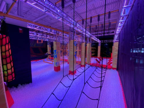 An indoor obstacle course with colorful lighting, featuring ropes, nets, and padded structures on a soft floor.