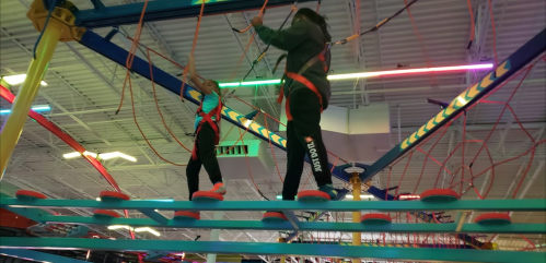 Two children navigate a colorful indoor ropes course, wearing safety harnesses and balancing on beams.