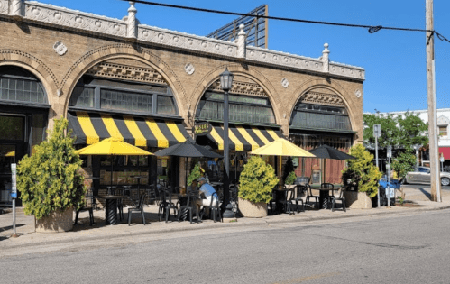 A café with yellow and black striped awnings, outdoor seating, and potted plants on a sunny day.
