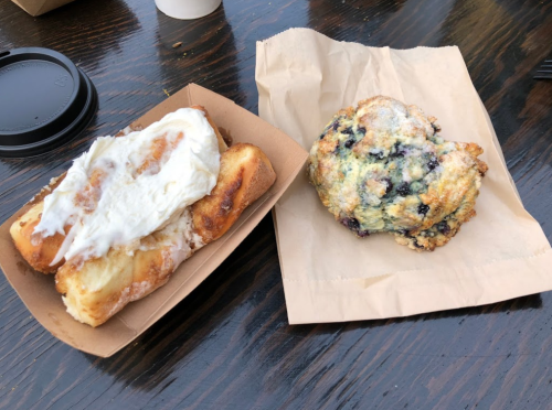 A cinnamon roll with icing next to a blueberry scone on a brown paper tray.
