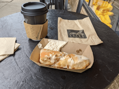 A coffee cup and a cinnamon roll on a table, with a napkin and a paper bag in the background.