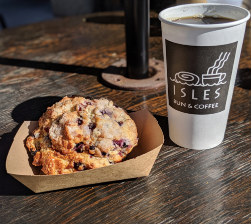 A blueberry muffin on a paper tray next to a cup of coffee from Isles Bun & Coffee on a wooden table.