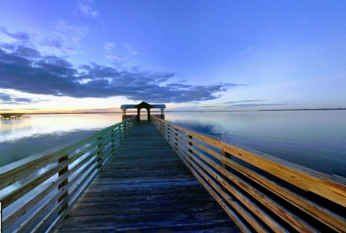 A wooden pier extends into calm waters under a colorful sky at dusk, with clouds reflecting on the surface.