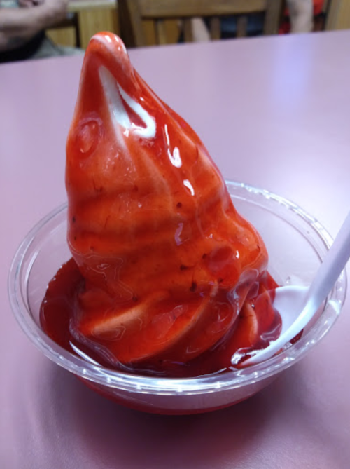 A cup of soft-serve ice cream swirled with red strawberry topping, served with a plastic spoon.