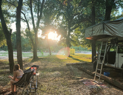 A person sits in a camping chair by a river, with a tent and trees in the background, as the sun sets.
