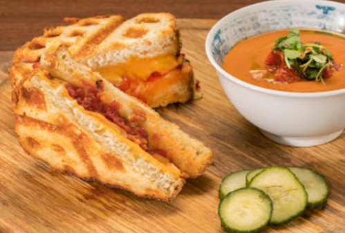 A grilled cheese sandwich with tomato and bacon, served with a bowl of tomato soup and cucumber slices on a wooden board.