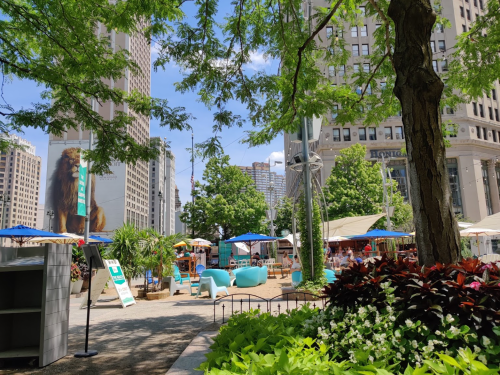 A vibrant urban park with seating, greenery, and colorful tents, surrounded by tall buildings on a sunny day.
