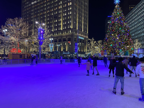A festive ice skating rink at night, surrounded by colorful holiday lights and a large Christmas tree.