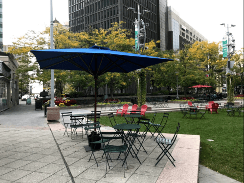 A park area with green grass, tables and chairs under a blue umbrella, surrounded by trees and buildings.