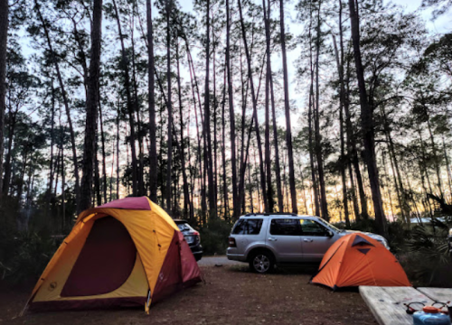 Two tents set up in a forested campsite with a parked car, surrounded by tall trees and a colorful sunset sky.