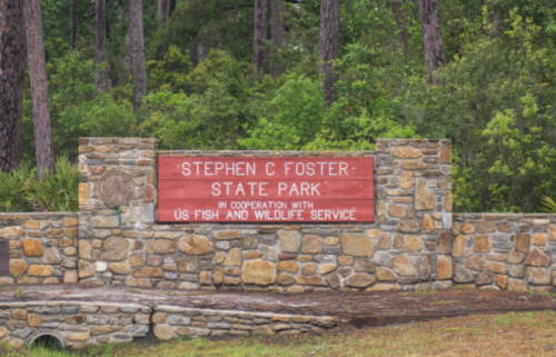 Sign for Stephen C. Foster State Park, featuring stone structure and surrounded by trees.