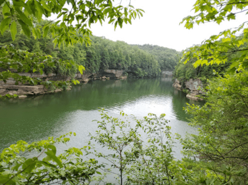 A serene lake surrounded by lush green trees and rocky cliffs under a cloudy sky.