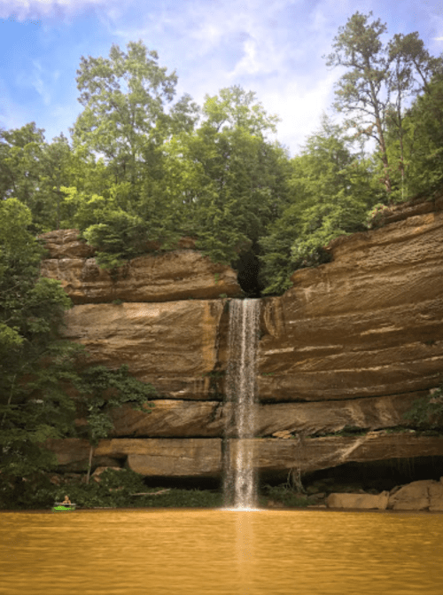 A serene waterfall cascades down rocky cliffs, surrounded by lush green trees and reflecting in a calm, muddy lake.
