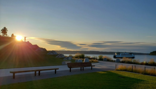 Sunset over a calm waterfront, with a ferry in the distance and a park featuring benches and grassy areas.
