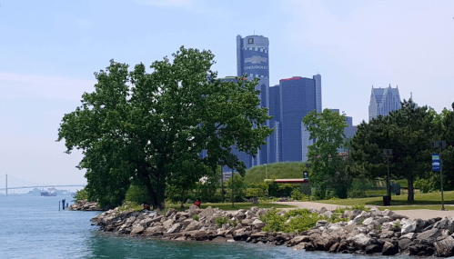 A scenic view of a waterfront park with trees, rocks, and city skyscrapers in the background on a sunny day.