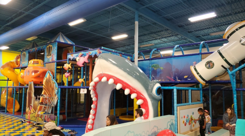 A colorful indoor playground featuring slides, a shark-shaped entrance, and children playing in various areas.