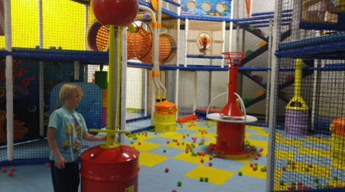 A child plays in a colorful indoor playground filled with climbing structures and scattered balls.