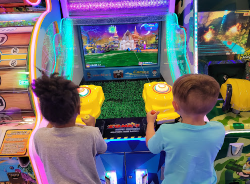 Two children playing a colorful arcade game with bright screens and joystick controls.