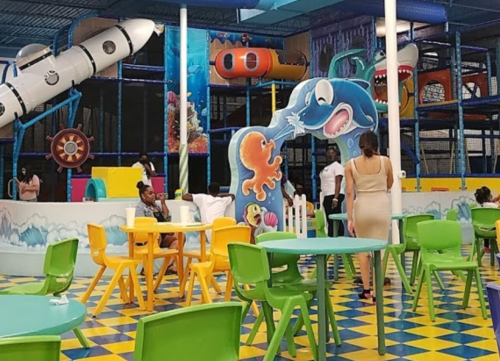 A colorful indoor play area with slides, climbing structures, and children playing at tables.