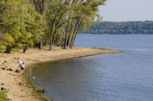A serene lakeside scene with sandy shore, trees, and people walking along the water's edge on a sunny day.