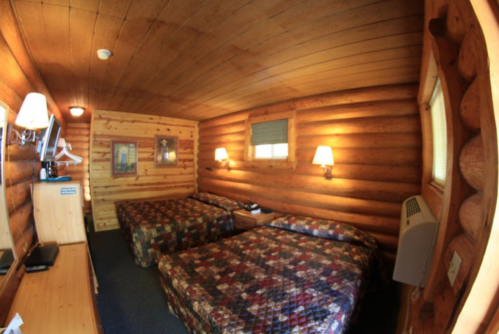 Cozy log cabin interior featuring two beds, wooden walls, lamps, and a small desk.