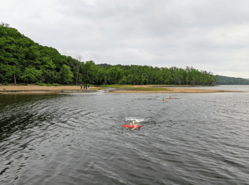 A calm river scene with swimmers in red life jackets and a green forested shore in the background.