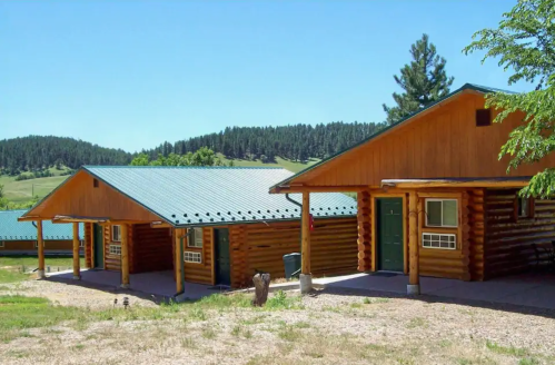 Two log cabins with green roofs set against a backdrop of rolling hills and trees under a clear blue sky.