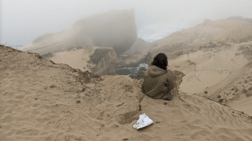 A person sits on a sandy cliff overlooking a foggy coastline, with a single shoe nearby.