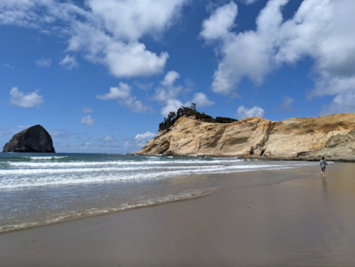 A person walks along a sandy beach with cliffs and a rock formation under a blue sky with fluffy clouds.