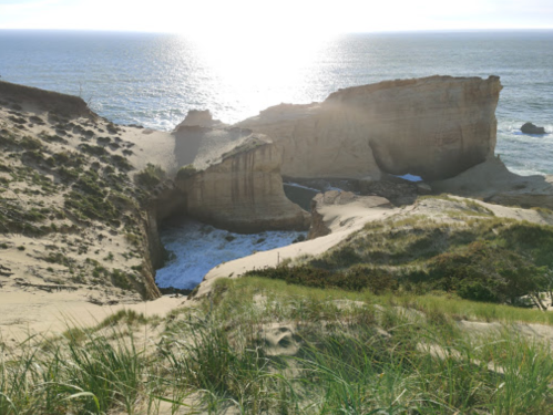 A coastal landscape featuring sandy cliffs, a rocky inlet, and sunlight reflecting on the ocean waves.