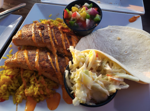A plate with two seasoned fish fillets, yellow rice, a side of coleslaw, and a fresh vegetable salsa in small bowls.
