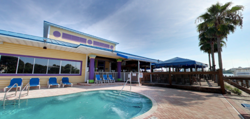 Colorful waterfront building with a pool, lounge chairs, and palm trees under a clear blue sky.