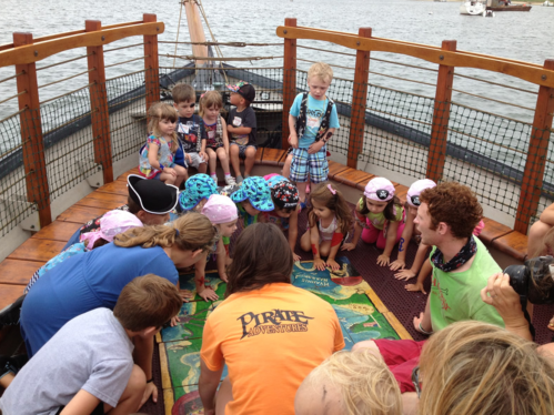 Children in pirate costumes gather around a map on a boat, engaged in an activity with adults.