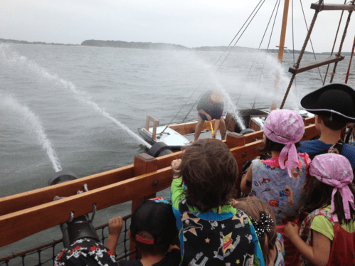 Children watch as a ship fires cannons into the water, creating splashes on a cloudy day.