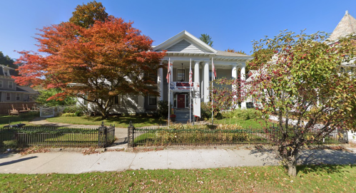 A large white house with a porch, surrounded by colorful autumn trees and a well-kept garden.