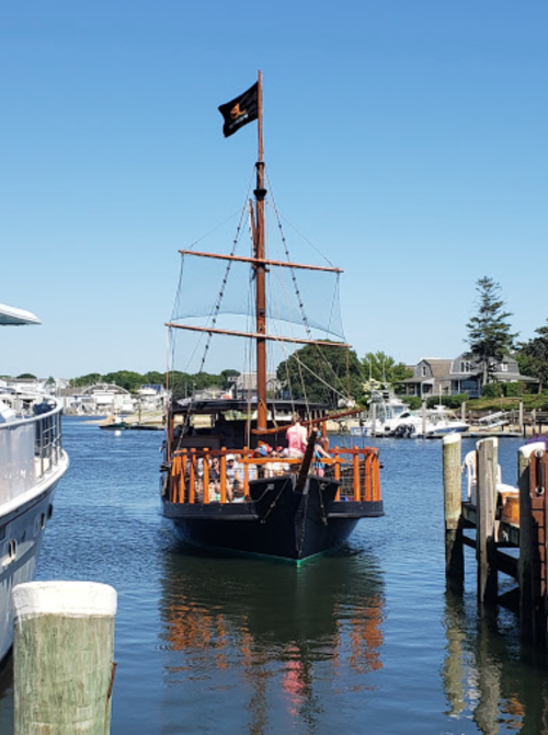 A wooden ship with a black flag sails through a marina on a clear, sunny day, surrounded by boats and homes.