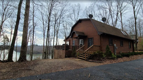 A log cabin by a lake, surrounded by bare trees and a gravel driveway, under a cloudy sky.