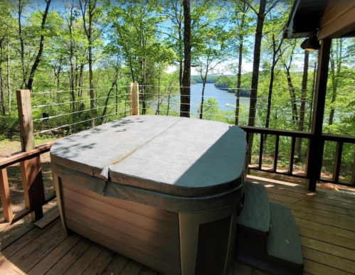 A hot tub on a wooden deck overlooking a serene lake surrounded by lush green trees.