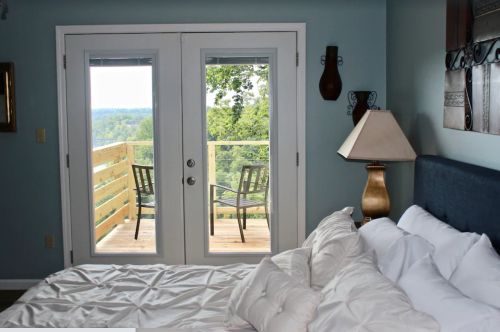 Cozy bedroom with a bed, lamp, and double doors leading to a balcony with two chairs and a scenic view.