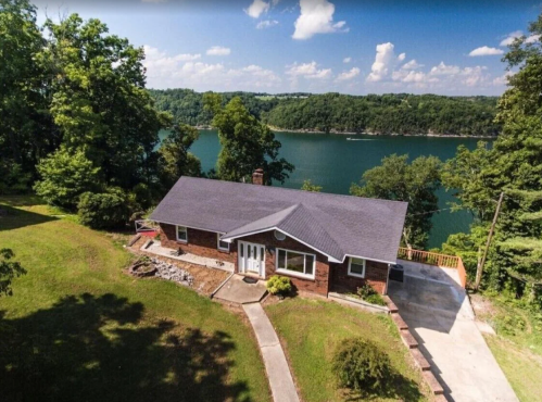 Aerial view of a brick house by a lake, surrounded by trees and greenery, with a clear blue sky above.