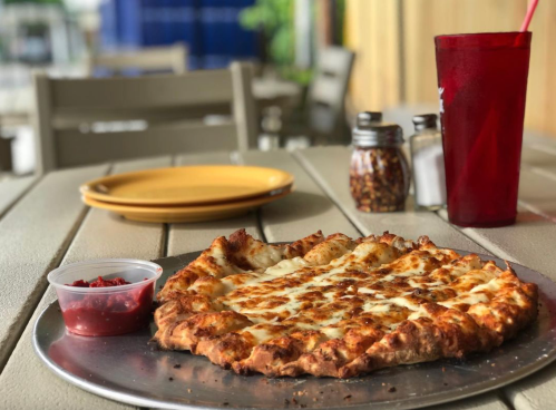 A freshly baked pizza on a metal tray, served with marinara sauce, on a table with plates and condiments in the background.