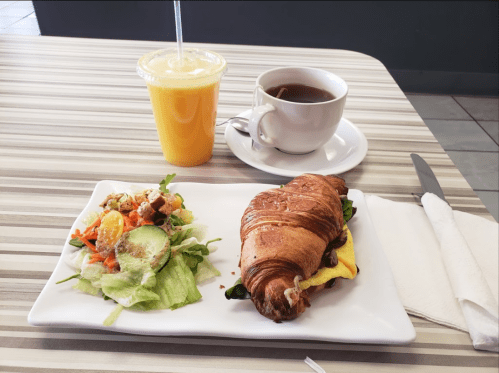 A croissant sandwich with salad, a cup of coffee, and a glass of orange juice on a striped table.