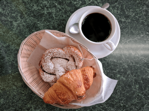 A basket with a powdered sugar pastry and a croissant, alongside a cup of black coffee on a green table.