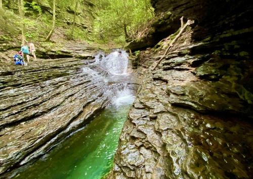 A serene waterfall cascades over layered rock formations in a lush green forest, with people exploring nearby.
