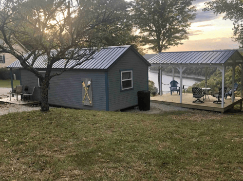 A small gray cabin with a metal roof beside a covered deck overlooking a lake, surrounded by trees.