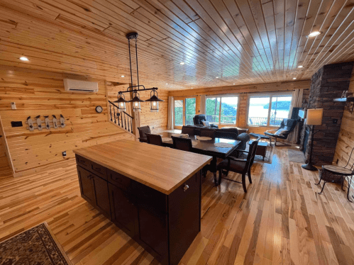 Cozy wooden interior of a spacious living area with a dining table, kitchen island, and large windows overlooking a lake.
