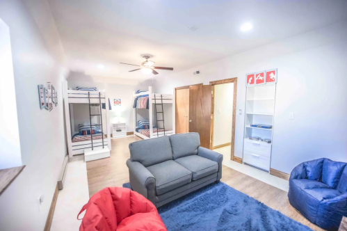 A cozy room with bunk beds, a gray sofa, bean bags, and a white shelving unit, featuring warm lighting and wooden accents.